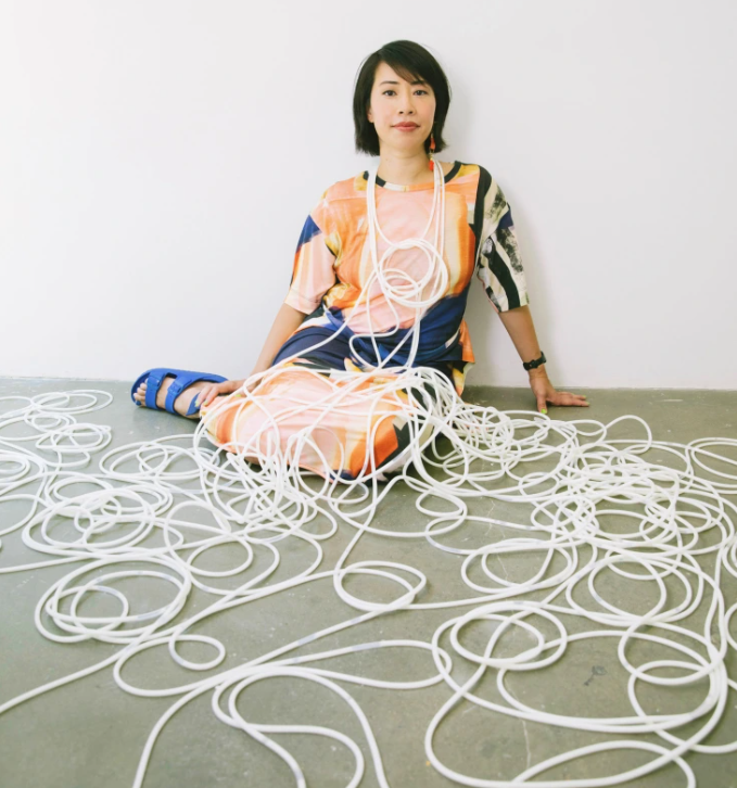 Portrait of Ani Liu sitting on the floor of Cuchifritos Gallery, covered in plastic tubes that have a white milk-like substance running through them. Photo taken by Celeste Sloman